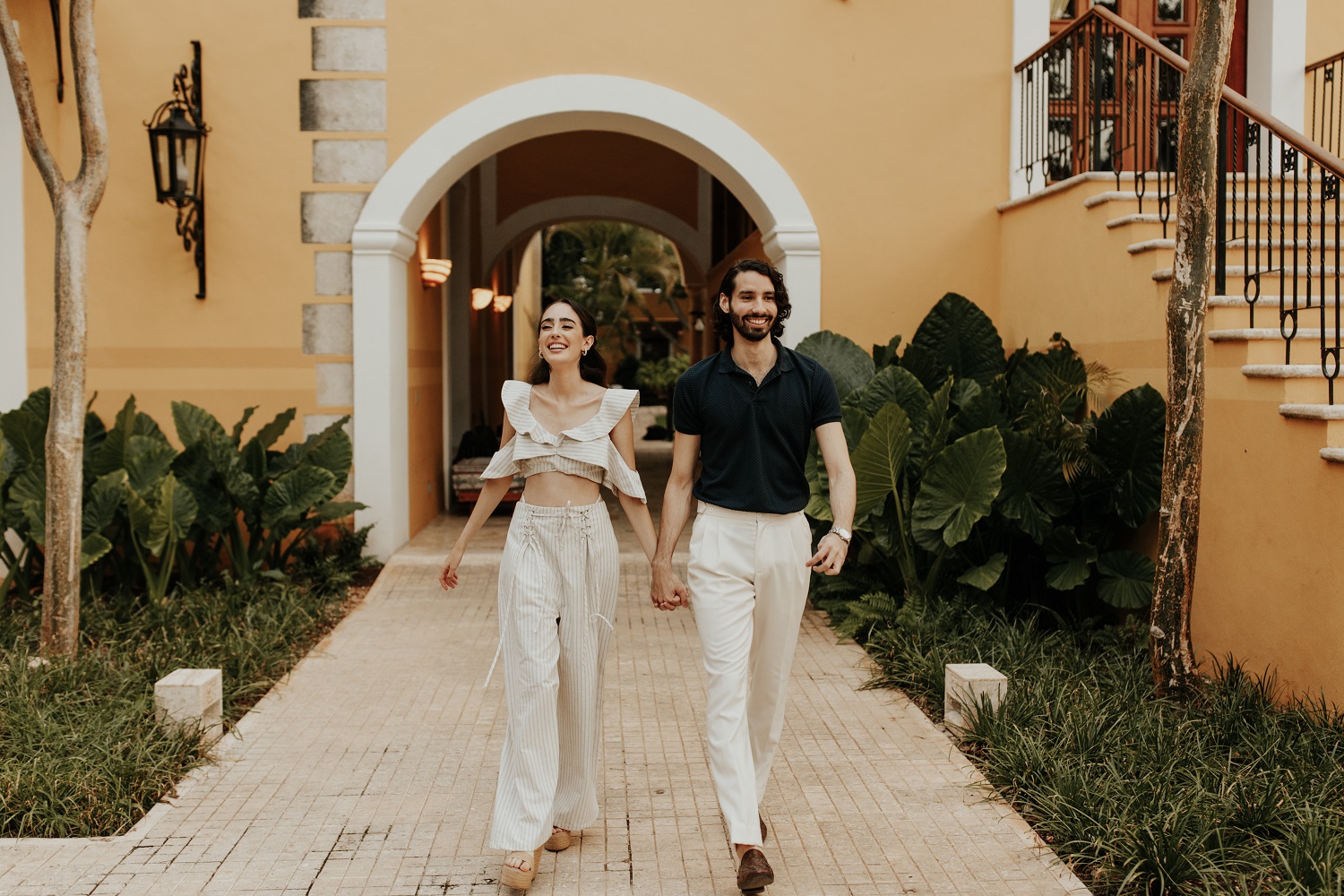 Couple walking hand-in-hand, smiling, in garden path of Hacienda Xcanatun, Angsana Heritage Collection.